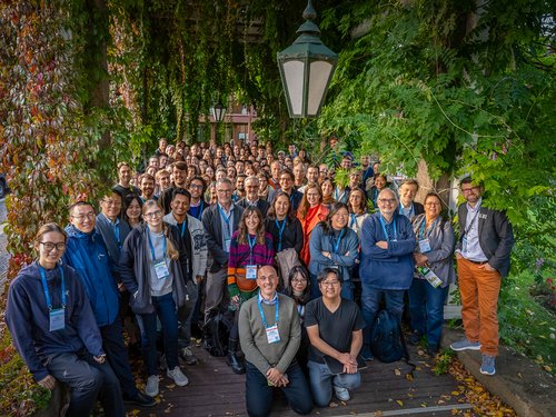 Große Gruppe von rund 200 Teilnehmenden der Learning & Memory Conference 2025 steht dicht beieinander im Grünen unter einer Pergola, lächelt in die Kamera. Im Vordergrund und Hintergrund sind Forschende verschiedener Nationalitäten zu sehen.
