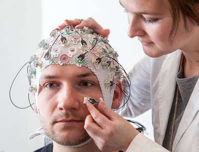 Wissenschaftlerin setzt Proband eine EEG-Haube auf und klebt Elektroden im Gesicht fest