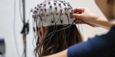 Forschende setzen einer Probandin eine EEG-Haube mit zahlreichen Elektroden auf, um Gehirnaktivität zu messen