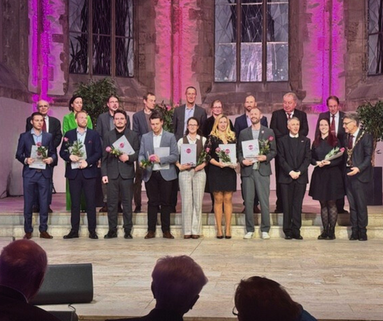 Gruppenfoto der Promotionspreisträgerinnen und -preisträger der Otto-von-Guericke-Universität Magdeburg beim Festakt in der Johanniskirche. Eine größere Gruppe steht auf einer Bühne vor beleuchteten Kirchenfenstern; alle halten Urkunden und Blumen in der Hand.