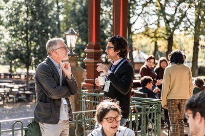 Mehrere Konferenzteilnehmende unterhalten sich bei Sonnenlicht und Kaffee auf einer Terrasse mit grünen Metallgeländern. Zwei Männer im Gespräch stehen im Vordergrund, im Hintergrund weitere Gruppen in lockerer Atmosphäre.