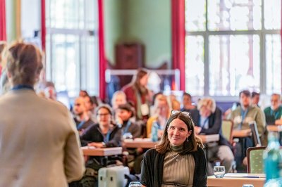 Teilnehmende der Konferenz sitzen in einem hellen, holzvertäfelten Saal und hören aufmerksam einer Vortragenden zu. Im Vordergrund ist eine Frau im Publikum fokussiert und lächelnd zu sehen.