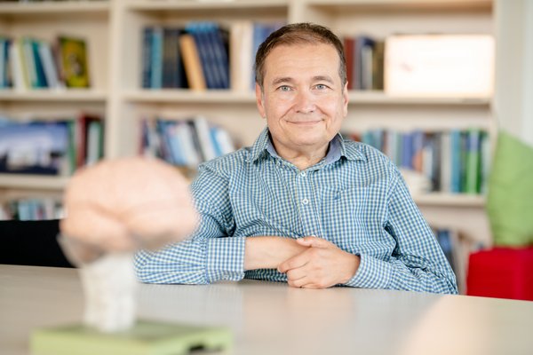 Ein Mann mittleren Alters im karierten Hemd sitzt an einem Tisch in einem Bücherzimmer und lächelt in die Kamera. Im Vordergrund unscharf ein Modell eines Gehirns.
