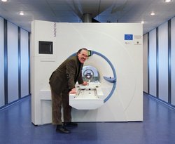Henning Scheich smiles while standing in front of a large MRI scanner, leaning on the patient table.